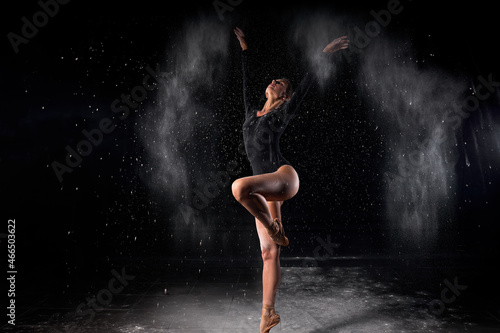 Artistic dance pose using powder or dust. Beautiful expressive ballet dancer un black swimsuit in dust at studio on black background.Slim caucasian Ballerina with flour. Full-length portrait