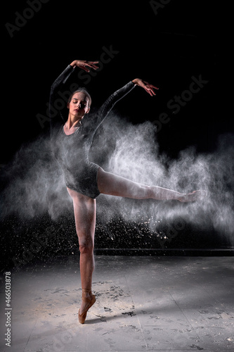 Dancer posing dancing gracefully in studio with cloud of dust  flour. Dancer in black bathing suit is moving  in action  having good choreographic training  raising leg up. ballet  dance  performance