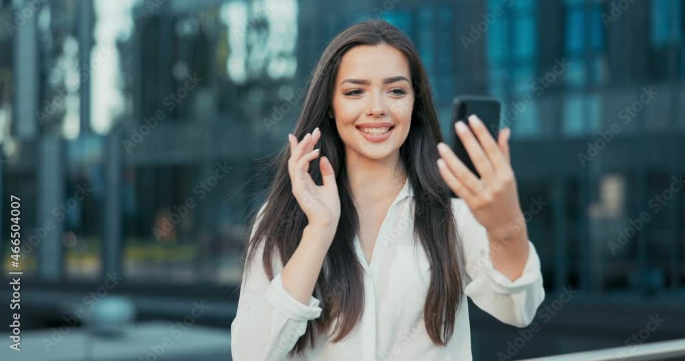 Smiling woman stands leaning against railing in front of modern glass office building, businesswoman dressed in white shirt holds phone in front of receives call waves hand to camera, video call Smiling woman stands leaning against railing in front of modern glass office building, businesswoman dressed in white shirt holds phone in front of receives call waves hand to camera, video call