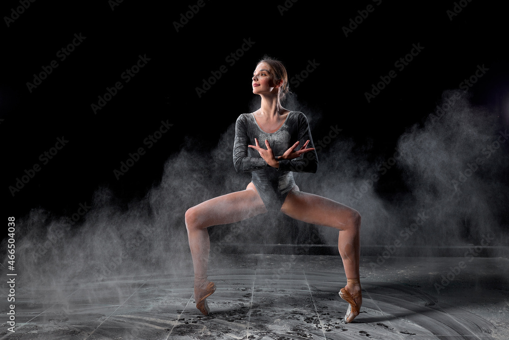 Flexible woman dancing with flour on black background in studio, moving with legs apart. Talented athlete ballet dancer in bodysuit in slow motion, looking at side, making dance tricks.