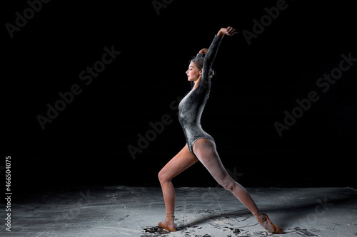 Dancing in flour concept. Slim beautiful female adult woman dancer in dust, fog. Lady is wearing black bodysuit, making dance element gracefully on isolated black background, side view