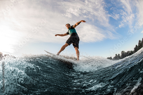 young man energetically balancing on wave on wakesurf board on splashing wave.