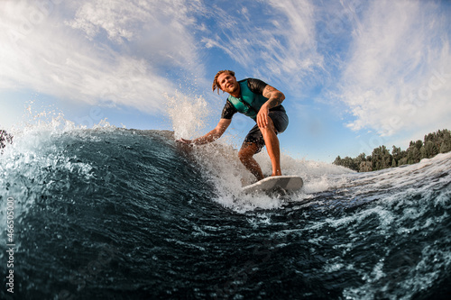 athletic man wakesurfer skilfully riding down the blue splashing wave on a warm day