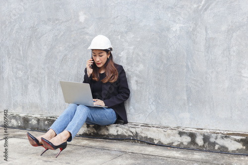 asian engineer developer woman wear black suit and white safety hardhat using laptop and talking mobile phone working onsite. foreman female work in construction. © eakgrungenerd