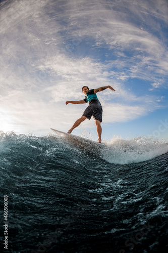 active man wakesurfer balancing on board on splashing river wave against blue sky