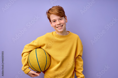 Happy teenage boy in yellow shirt holding basketball ball in hands isolated in studio on purple background, portrait. pozitive excited kid looking at camera, laughing, sport, childhood concept photo