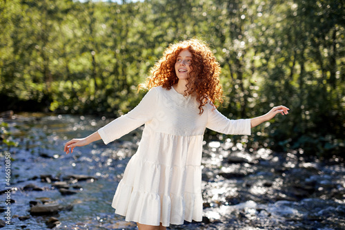 Happy sorceress redhead woman stands in white dress near river which flows in the mountain forest on sumemr sunny day, enjoy nature, looking around, having natural red curly hair. portrait photo