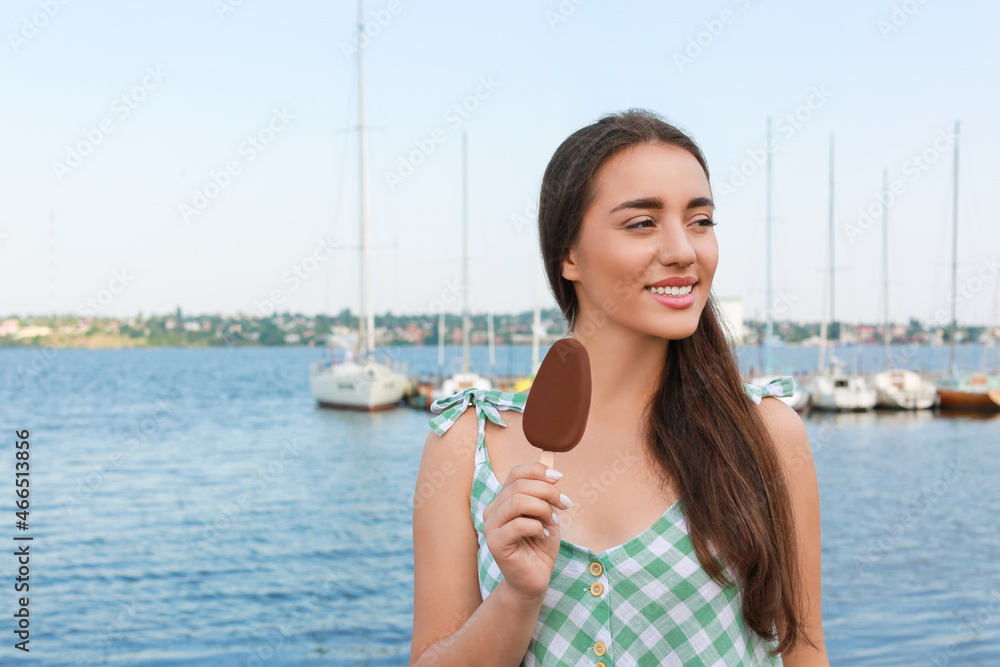 Fototapeta premium Beautiful young woman holding ice cream glazed in chocolate near river, space for text