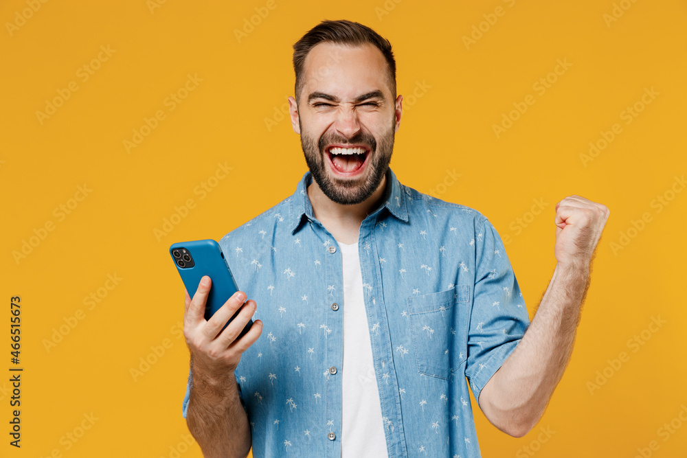 Young overjoyed cool happy caucasian man 20s wear blue shirt white t-shirt hold in hand use mobile cell phone chatting surfing do winner gesture isolated on plain yellow background studio portrait - obrazy, fototapety, plakaty 