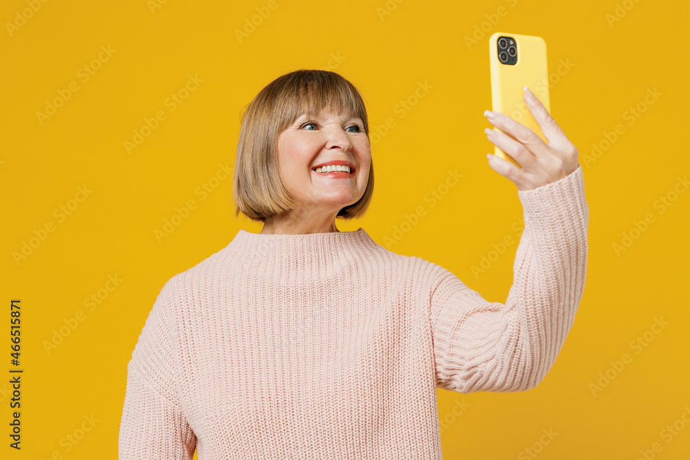 Elderly woman 50s wears pink casual knitted sweater doing selfie shot on mobile cell phone post photo on social network isolated on plain yellow background studio portrait. People lifestyle concept. Elderly woman 50s wears pink casual knitted sweater doing selfie shot on mobile cell phone post photo on social network isolated on plain yellow background studio portrait. People lifestyle concept.