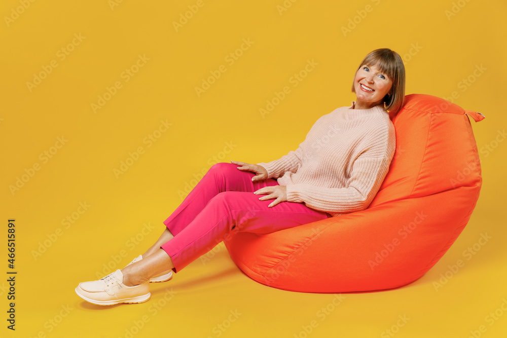 Full body smiling happy cheerful elderly caucasian woman 50s wear pink casual knitted sweater look camera sit in bag chair isolated on plain yellow background studio portrait People lifestyle concept