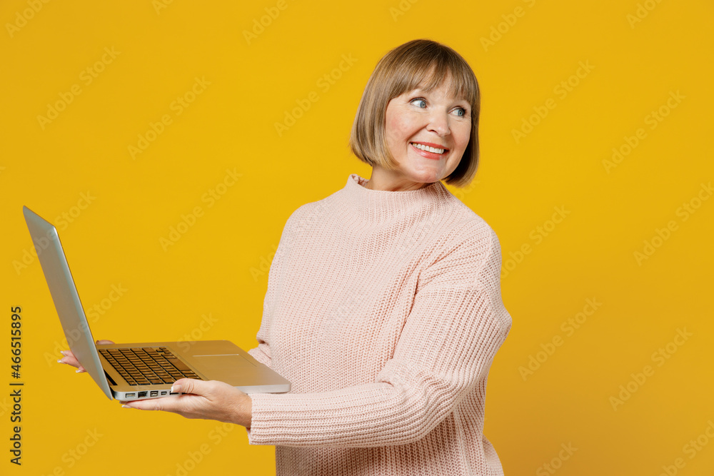 Naklejka premium Elderly smiling happy caucasian copywriter woman 50s wears pink casual knitted sweater hold use work on laptop pc computer look aside on copy space isolated on plain yellow background studio portrait