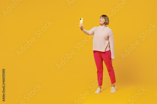 Elderly fun woman 50s wears pink casual knitted sweater do selfie shot on mobile cell phone post photo on social network isolated on plain yellow background studio portrait. People lifestyle concept.