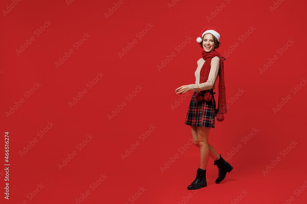 Full body side view young smiling caucasian woman 20s wear Santa Claus Christmas red hat walk going strolling isolated on plain red background studio portrait. Happy New Year 2022 celebration concept.