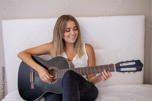 Chica sentada en la cama tocando la guitarra. Chica sonriendo. Mujer rubia practicando un hobbie. Disfrutar de la música. Instrumento de cuerda. Guitarra negra.