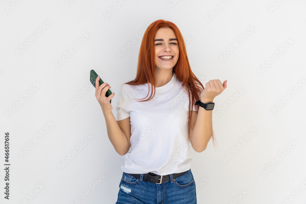 Portrait of redhead woman celebrating, shouting yes with satisfaction, fist pump, holding smartphone, winning on mobile phone, achieve daily goal in application over white background Portrait of redhead woman celebrating, shouting yes with satisfaction, fist pump, holding smartphone, winning on mobile phone, achieve daily goal in application over white background