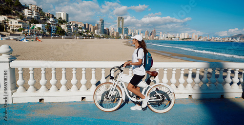 Happy woman tourist with electric bicycle over background Levante beach overlooking panorama of Benidorm city. Traveler concept photo