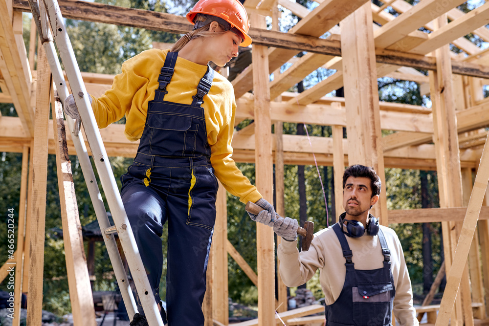 confident male in uniform helps young caucasian female constructor standing in height on ladder, in hardhat, bearded responsive male is giving hammer, working as team. friendly and cooperative.