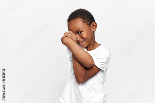 Shy and charming little preschool male kid covering face with awkwardly crossed arms, smiling with embarrassment, peeking between hands with one eye. Children and their emotions. Body language