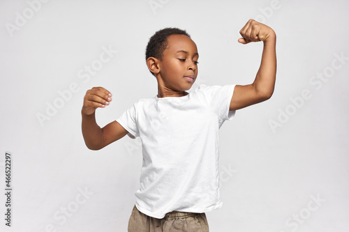 Portrait of strong confident dark-skinned boy, contracting upper arm muscles, showing biceps and triceps after doing workout, dressed in white sport t-shirt, feeling proud, self-satisfied