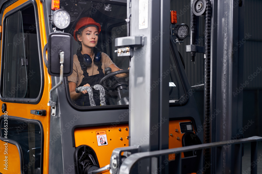 focused Female worker driving loader in industrial container warehouse, looking confident, dressed in engineer uniform and hardhat orange helmet, concentrated on work. agriculture concept