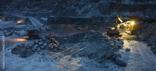 Quarry excavator and quarry dump trucks in the process of loading stone ore in a limestone quarry during night work in the winter, with artificial lighting, image in blue cold tones, panorama.