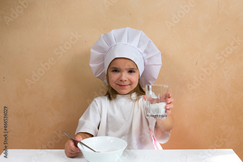 Little cheerful smiling girl dressed as a cook shows a glass of flour for kneading dough.