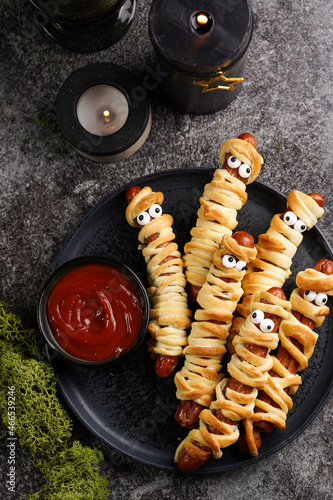 Cute Halloween party snack: wiener sausage with sugar eyes wrapped in dough stripes resembling mummies on a black plate with tomato ketchup