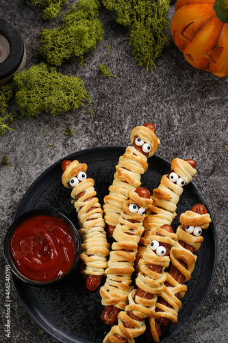 Cute Halloween party snack: wiener sausage with sugar eyes wrapped in dough stripes resembling mummies on a black plate with tomato ketchup