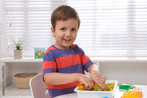 Cute little boy playing with bright kinetic sand at table in room