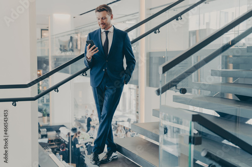 Stunning entrepreneur in formal stylish suit standing on staircase in office lobby with smartphone