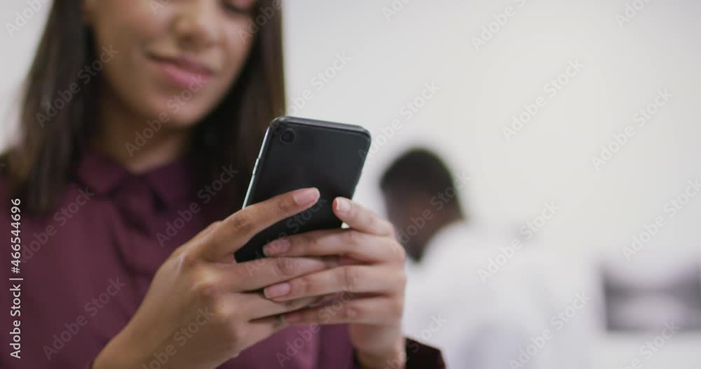 Smiling biracial businesswoman using smartphone with colleagues in background in modern office Smiling biracial businesswoman using smartphone with colleagues in background in modern office