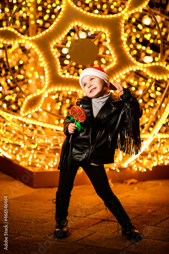 a beautiful, cheerful little girl in a Santa Claus hat,standing near the lights,with a lollipop in her hand on a festive evening, on the street