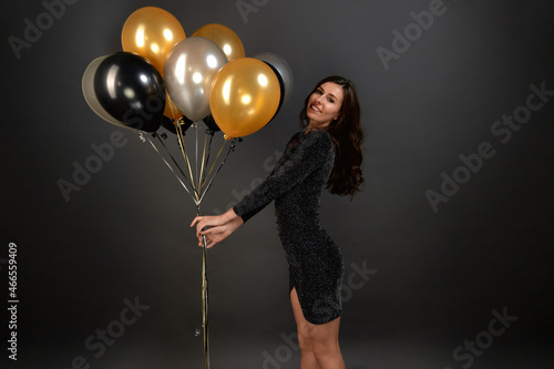 Smiling woman with dark long hair holds gold and silver balloons, preparing surprise for new year party, celebrates holiday, wears sparkly evening sequin dress on black background. Festive concept.