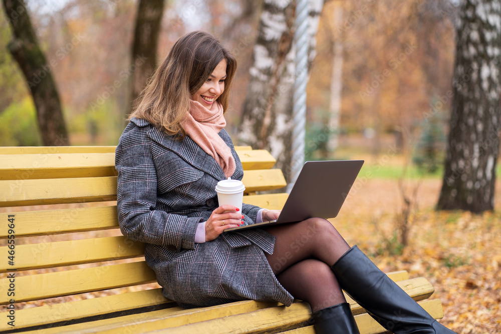 Girl sits on a park bench in autumn with a laptop and drinks coffee Girl sits on a park bench in autumn with a laptop and drinks coffee