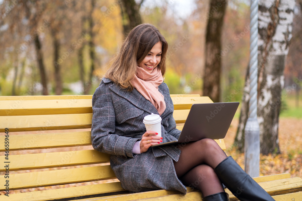 Girl sits on a park bench in autumn with a laptop and drinks coffee Girl sits on a park bench in autumn with a laptop and drinks coffee