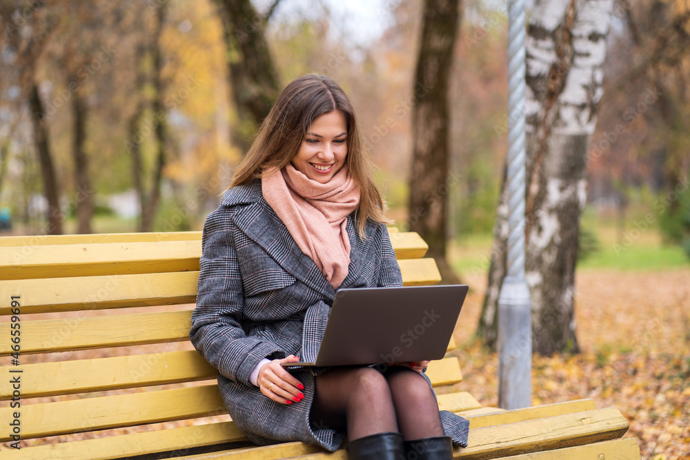Girl sits on a park bench in autumn with a laptop and drinks coffee Girl sits on a park bench in autumn with a laptop and drinks coffee