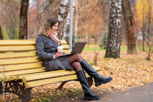 Girl sits on a park bench in autumn with a laptop and drinks coffee