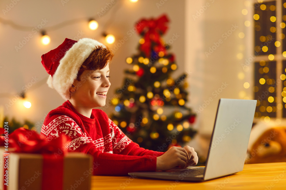 Christmas Eve at home. Little boy in a Santa hat is sitting in front of a laptop and talking on a video call. Child spends the Christmas holidays at home communicating with everyone over the internet. Christmas Eve at home. Little boy in a Santa hat is sitting in front of a laptop and talking on a video call. Child spends the Christmas holidays at home communicating with everyone over the internet.
