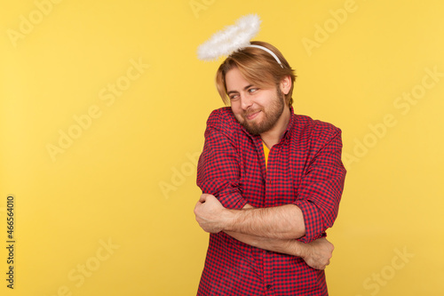 Portrait of hipster bearded guy in checkered shirt and with nimbus overhead hugging himself with love, worship to beauty, over-inflated ego. Indoor studio shot isolated on yellow background.