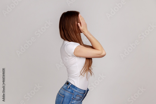 Side view portrait of young adult dark haired woman standing, covering eyes with hand, refusing to watch, shame content, wearing white T-shirt. Indoor studio shot isolated on gray background.