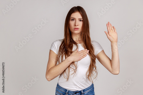 Portrait of very responsible and honest woman giving promise, making solemn vow in ceremonial tradition with raised hand, wearing white T-shirt. Indoor studio shot isolated on gray background.