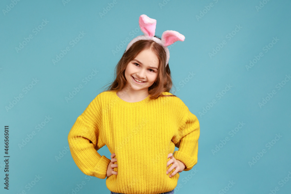Portrait of cute little girl stands with pink bunny ears, child having fun, keeping hands on hips, wearing yellow casual style sweater. Indoor studio shot isolated on blue background. Portrait of cute little girl stands with pink bunny ears, child having fun, keeping hands on hips, wearing yellow casual style sweater. Indoor studio shot isolated on blue background.