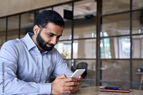 Smiling indian ceo businessman holding using cell phone mobile apps sitting at office desk looking at screen. Digital tech apps and solutions for business corporate development.