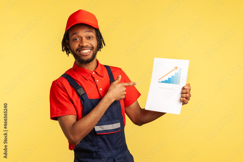 Portrait of smiling satisfied workman with beard wearing blue uniform, holding paper with graph showing financial increase of service industry. Indoor studio shot isolated on yellow background.