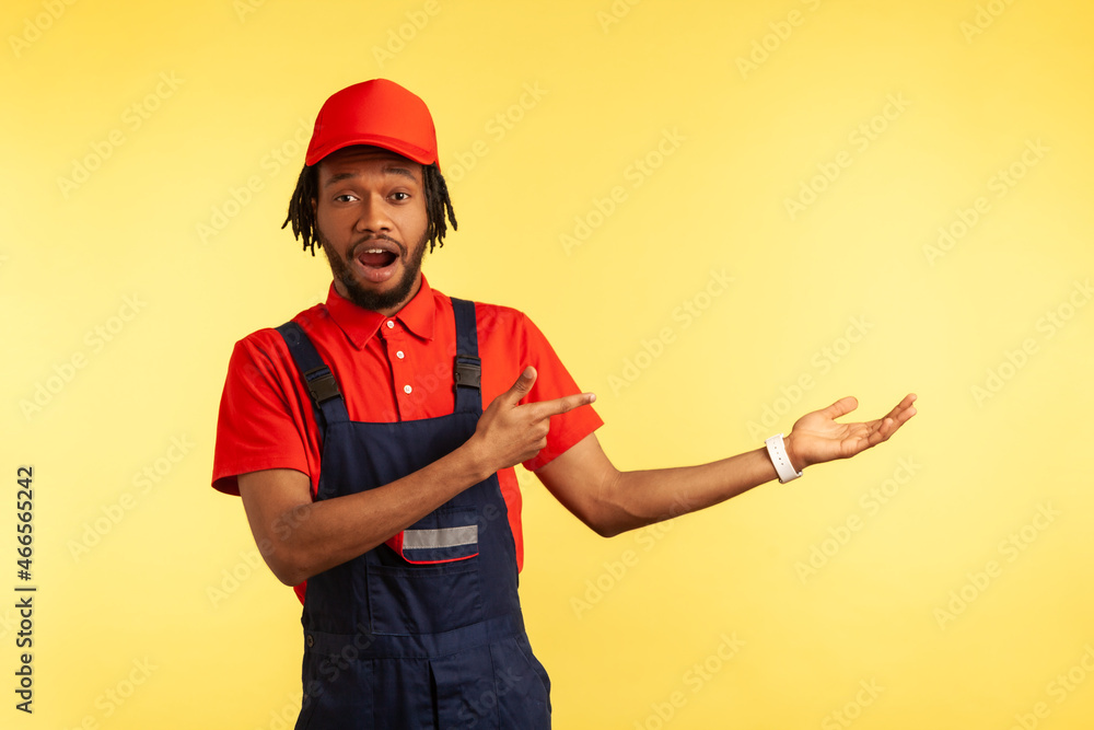Astonished builder or courier wearing overalls, presenting copy space for advertisement, pointing finger aside, ad of delivery service. Indoor studio shot isolated on yellow background.