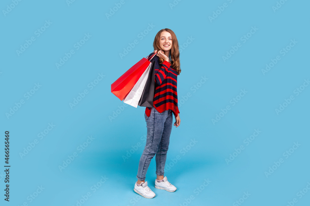 Full length portrait of beautiful woman wearing striped casual style sweater, standing holding shopping bags or gift and looking at camera with smile. Indoor studio shot isolated on blue background. Full length portrait of beautiful woman wearing striped casual style sweater, standing holding shopping bags or gift and looking at camera with smile. Indoor studio shot isolated on blue background.