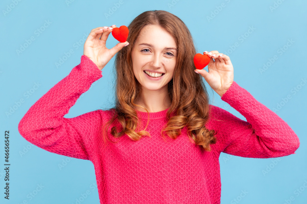 Portrait of optimistic young adult woman wearing pink pullover, having fun with red toy hearts, showing his fondness and devotion, romance. Indoor studio shot isolated on blue background. Portrait of optimistic young adult woman wearing pink pullover, having fun with red toy hearts, showing his fondness and devotion, romance. Indoor studio shot isolated on blue background.