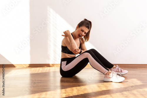 Girl touches painful elbow, suffers ulnar joint injury during training, sport trauma, inflamed nerve, wearing black sports top and tights. Full length studio shot illuminated by sunlight from window.