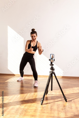 Fitness vlogger recording tutorials for sporty class, showing thumbs up to phone camera on tripod, wearing black sports top and tights. Full length studio shot illuminated by sunlight from window.